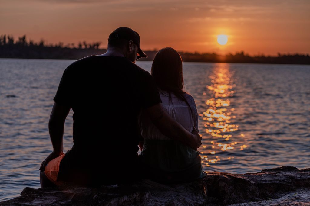 A couple sitting close together on a rock by the lake at sunset, enjoying the serene view.